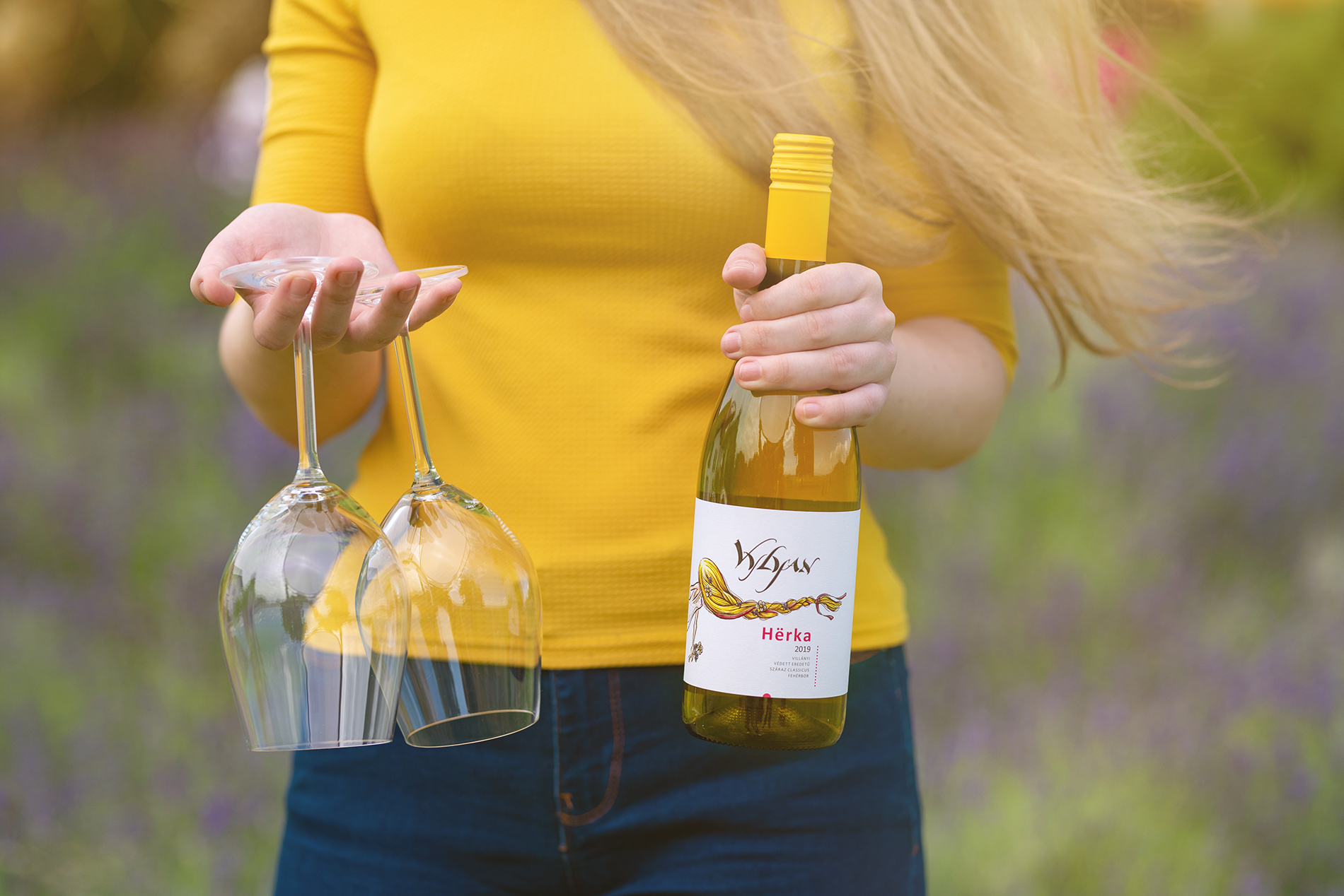 A kezében két poharat és 2019-es Vylyan Herkat tartó lány a szélben, a Vylyan birtokon. 